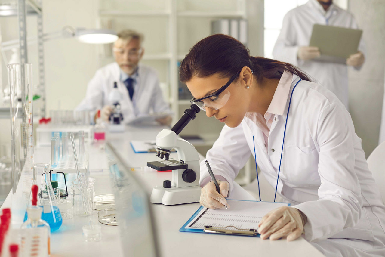 Serious scientist in lab coat, goggles and gloves doing advanced research, conducting laboratory experiment, looking in optical microscope, collecting data, taking notes on clipboard, writing report